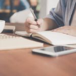 Scholarship Application - Woman Writing on a Notebook Beside Teacup and Tablet Computer