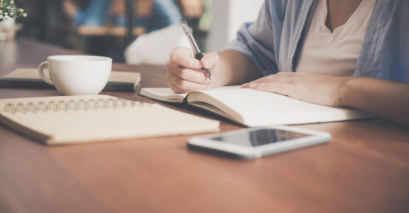 Scholarship Application - Woman Writing on a Notebook Beside Teacup and Tablet Computer