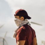 Sustainable Travel - Unrecognizable man with scarf on face walking in sandy terrain terrain near windmills