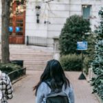 Education Path - Full body back view females with backpacks wearing casual outfits strolling together on paved walkway towards modern building