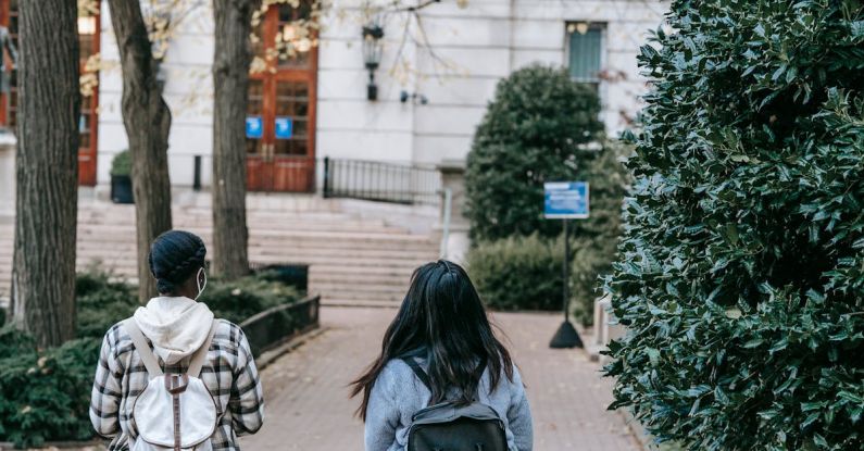 Education Path - Full body back view females with backpacks wearing casual outfits strolling together on paved walkway towards modern building